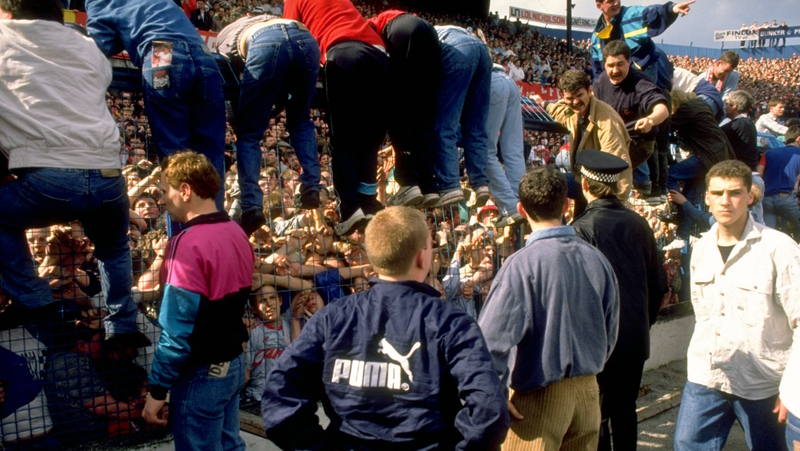 Fans climb over the steel barrier surrounding the pitch to avoid being crushed at Hillsborough Stadium. (Photo via David Cannnon/Getty).